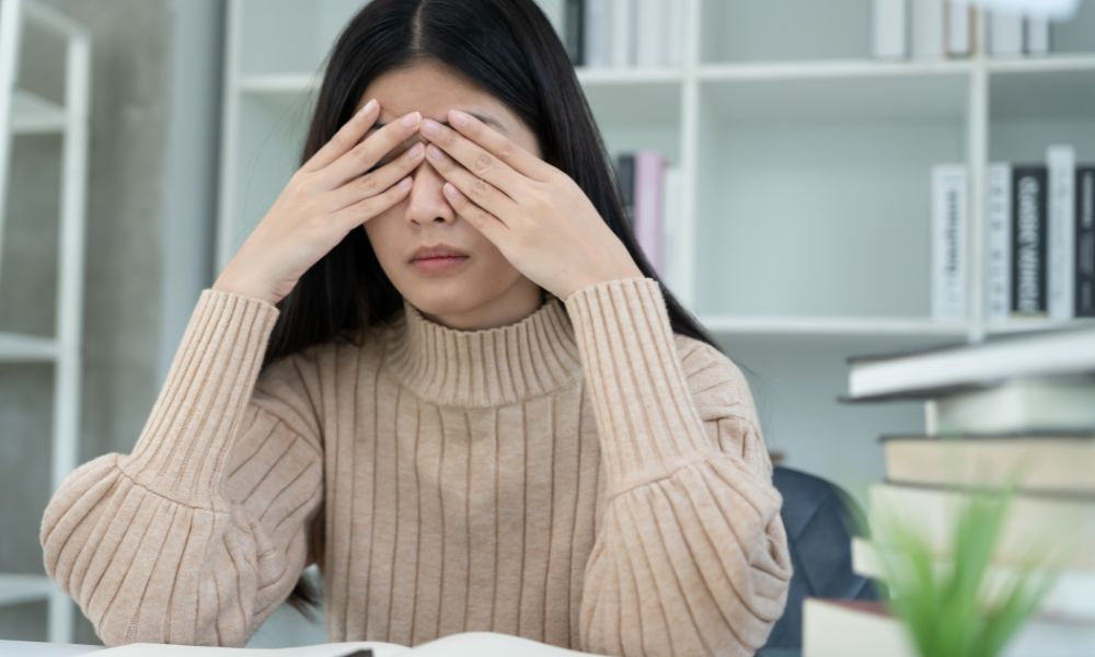  Žena si zakrýva oči rukami pri práci s knihami, čo poukazuje na unavené oči, digitálnu únavu zraku a nepríjemné pálenie či rezanie očí počas dňa.