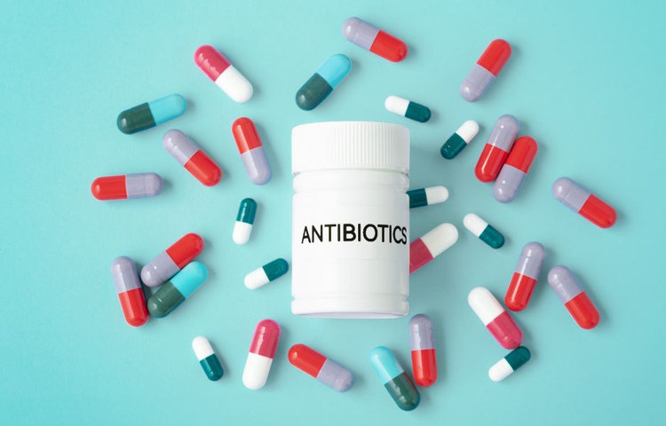 Biela fľaša s nápisom Antibiotics obklopená farebnými kapsulami rôznych farieb na tyrkysovom pozadí, znázorňujúca potrebu užívať probiotiká po antibiotikách na obnovu črevného mikrobiómu.
