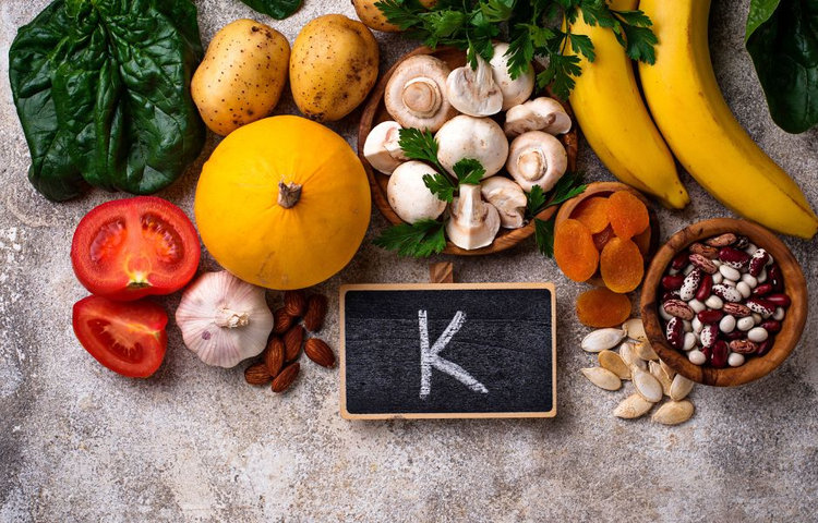 Pohľad zhora na pestrofarebné potraviny bohaté na draslík a kalium vrátane banánov, húb, paradajok, špenátu a orechov s tabuľkou s nápisom K ako symbol minerálu.