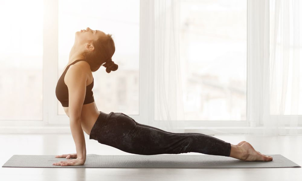  Žena v športovom oblečení vykonáva jogovú pozíciu kobry na podložke pri veľkom okne, ktorá patrí medzi odporúčané ischias cviky na uvoľnenie zápal sedacieho nervu.