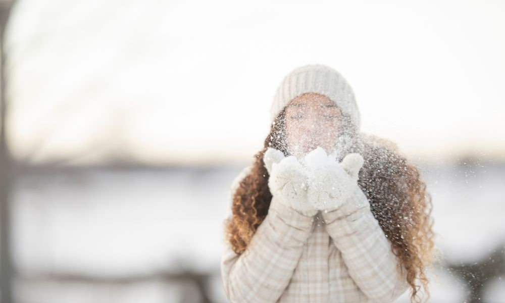  Žena v zimnom oblečení fúka sneh z dlaní, čo pripomína potrebu zvýšenej starostlivosti o pokožku v decembri, najmä hydratácie, ochrany pred mrazom a pravidelného čistenia pleti.