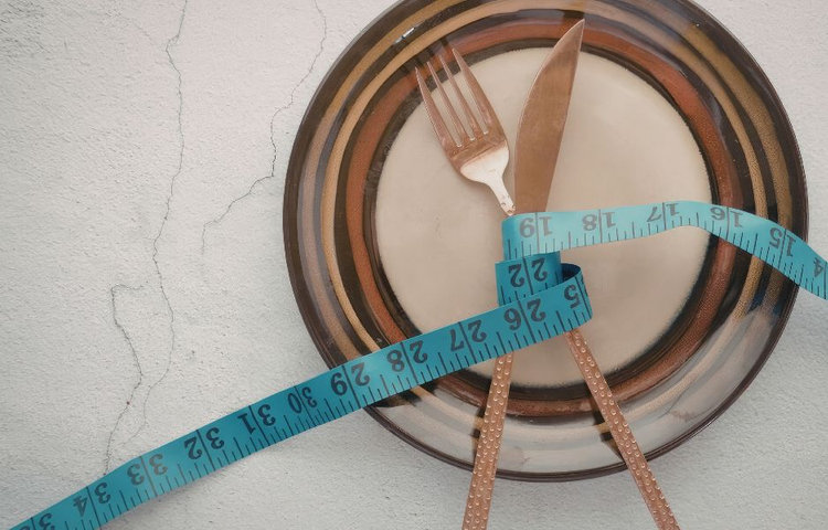 Prázdny tanier s vidličkou a nožom previazanými modrým krajčírskym metrom na svetlom pozadí, symbolizujúci kontrolu stravy a žlčníkovú diétu zameranú na to, čo jesť pri žlčníku.