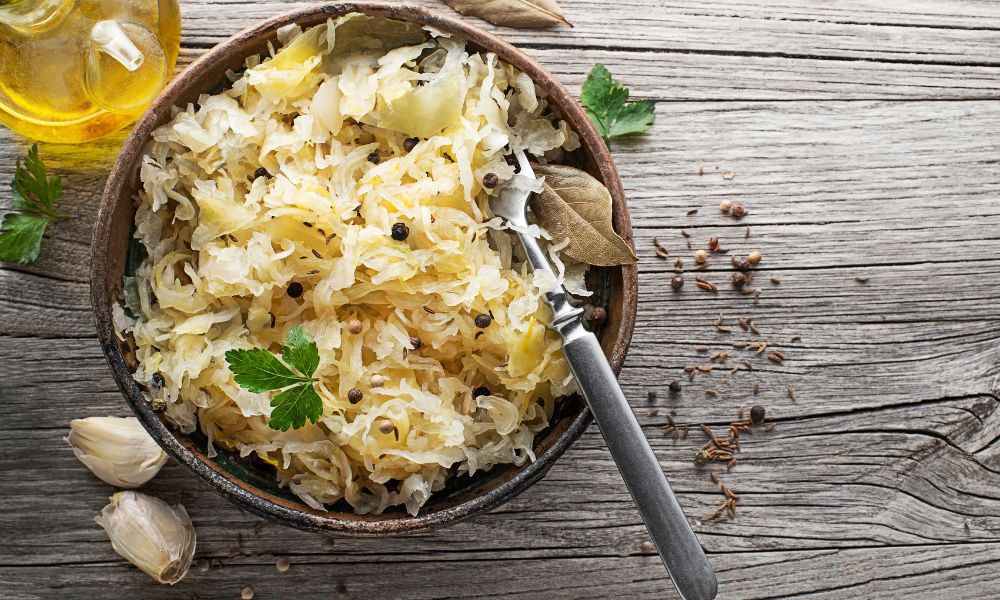  Miska kyslej kapusty s vidličkou, cesnakom a korením na drevenom povrchu vedľa fľaše oleja, zobrazujúca fermentované potraviny bohaté na probiotiká prospešné pre črevný mikrobióm.