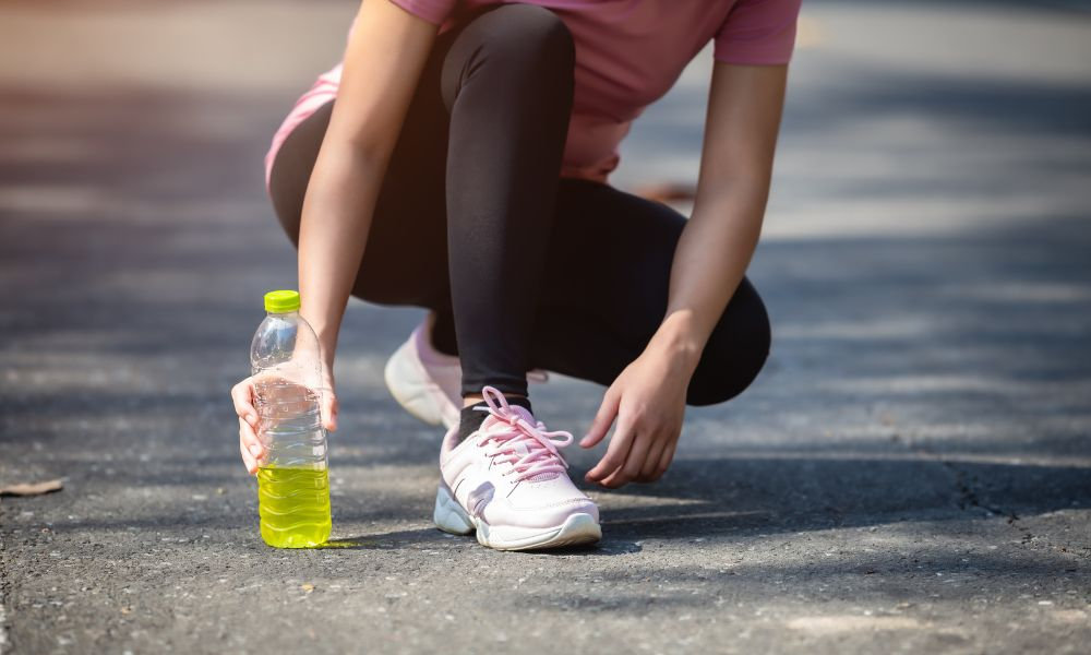  Športovkyňa v ružovom oblečení si zaväzuje tenisky a drží fľašu so žltým elektrolytovým nápojom na asfaltovej ceste pred behom vonku.