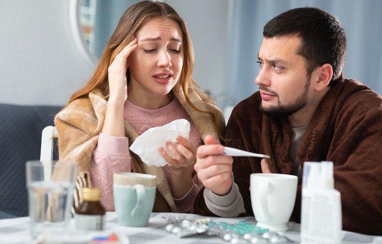 Muž sa stará o chorú ženu, ktorá má chrípku a drží v ruke teplomer.