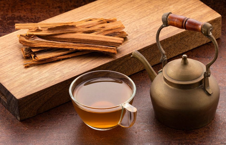 Sklenená šálka s čajom z vilcacory vedľa starej mosadznej kanvice a kusov sušenej kôry mačacieho pazúra na drevenej doske, zobrazujúca prípravu vilcacora čaju.