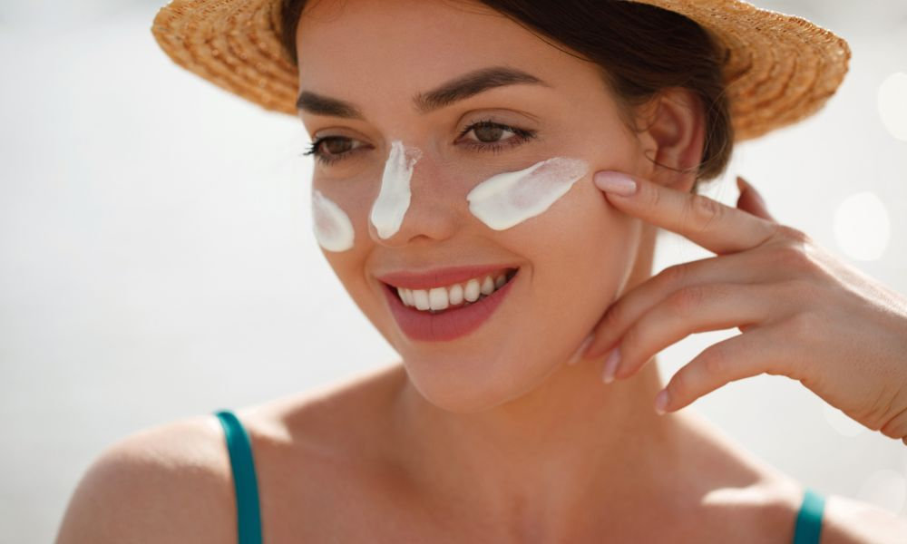  Usmiata žena so slamením klobúkom si nanáša opaľovací krém na tvár vo forme bielych pruhov, zobrazujúca ochranu pleti pred UVA žiarením pomocou opaľovacieho krému na tvár.