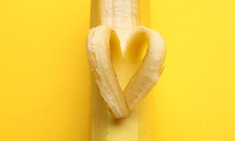  Ošúpaný banán so šupkou stočenou do tvaru srdca na žltom pozadí, symbolizujúci zdravie a lásku k potravinám bohatým na draslík a kalium.