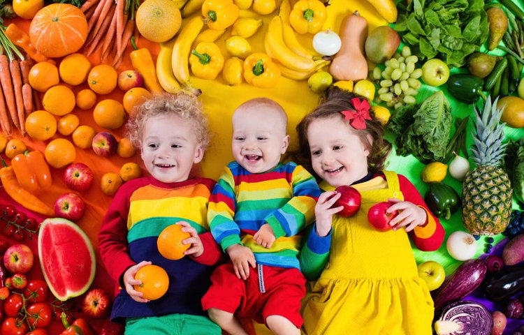 Tri malé deti ležia medzi pestrým ovocím a zeleninou, usmievajú sa a držia ovocie v rukách, čo symbolizuje zdravú stravu a význam probiotík pre deti a podporu imunity.