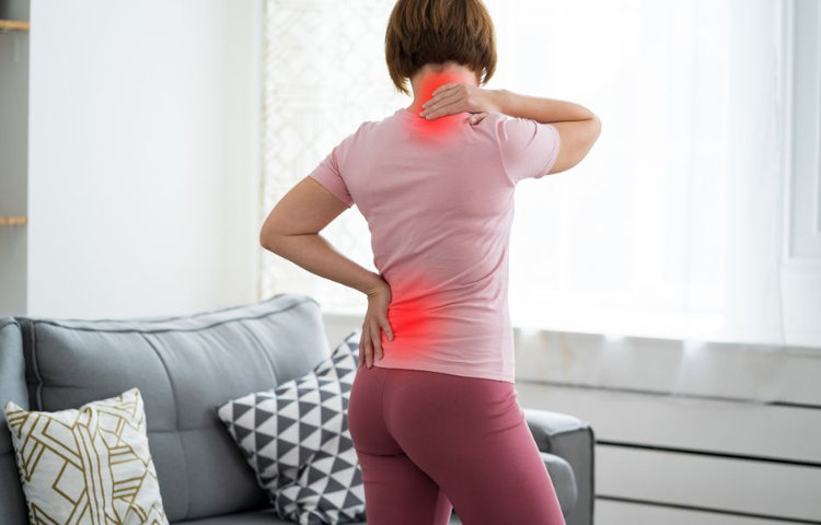 Žena v ružovom tričku stojí chrbtom a drží si ruky na bolestivých miestach krku a driekovej oblasti chrbta, ktoré sú graficky zvýraznené červenou farbou ako príznaky ischiasu.