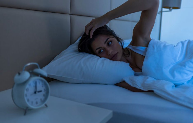 Žena leží v noci v posteli s rukou na hlave a bdelo hľadí pred seba na budík, čo zobrazuje nespavosť a narušený spánok ako typický príznak spánkového apnoe.