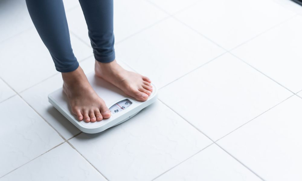 Bosé nohy osoby v športových nohaviciach stojace na bielej osobnej váhe na dlažbe, čo súvisí s udržiavaním zdravej hmotnosti ako súčasťou žlčníkovej diéty.
