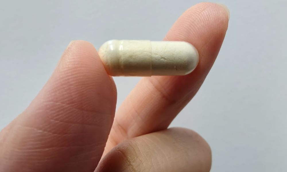  Ruka drží medzi prstami jednu bielu probiotickú kapsulu na svetlom pozadí, zobrazujúca najlepšie probiotiká k antibiotikám vo forme doplnku výživy.