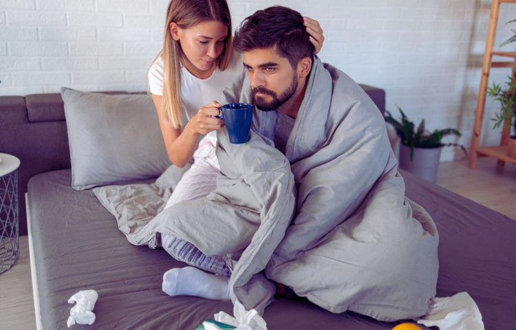 Muž zabalený v deke sediaci na posteli. Vedľa neho je žena, ktorá mu podáva šálku, pričom na zemi sú rozhádzané použité vreckovky, čo naznačuje, že je chorý a ona sa o neho stará.