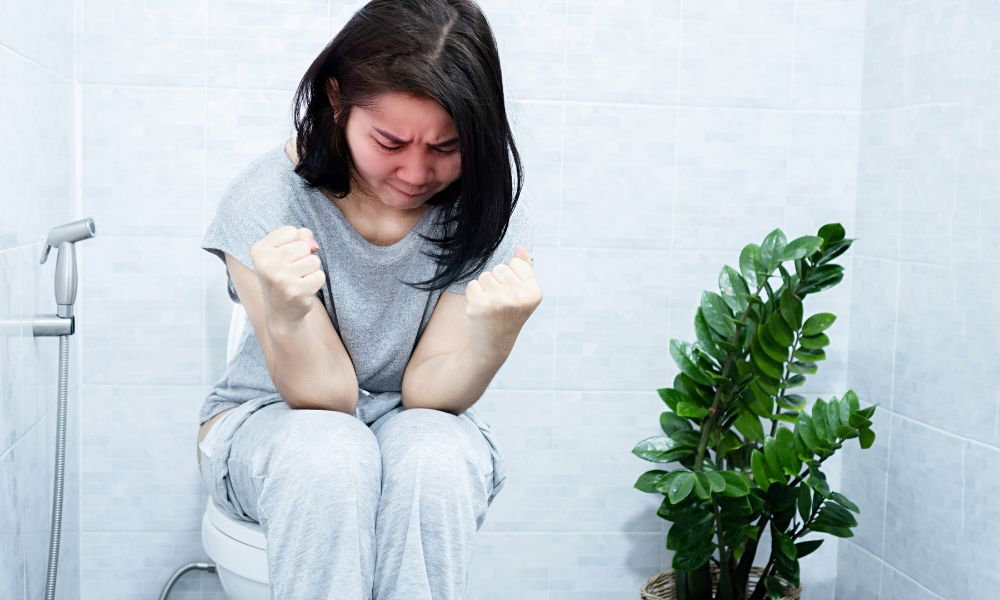  Žena sedí na toalete so zatnutými päsťami a bolesťou brucha, čo vystihuje nepríjemné prejavy zápchy u seniorov aj mladších ľudí pri dlhodobých tráviacich ťažkostiach.
