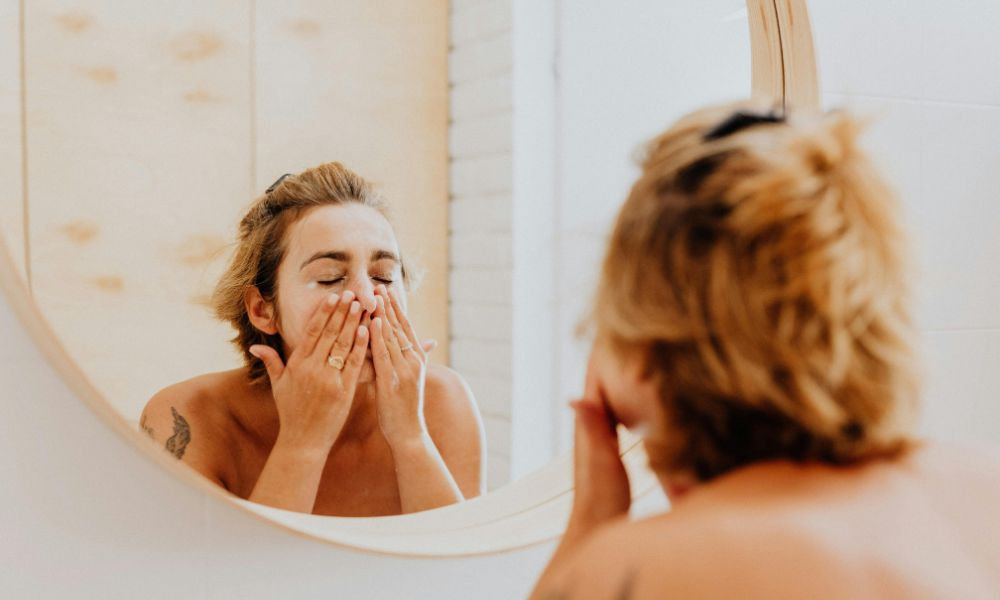  Žena si pri kúpeľňovom zrkadle umýva tvár čistiacim prípravkom, čo symbolizuje dôležitosť každodenného čistenia pleti a prípravy pokožky na hydratáciu počas zimných mesiacov.
