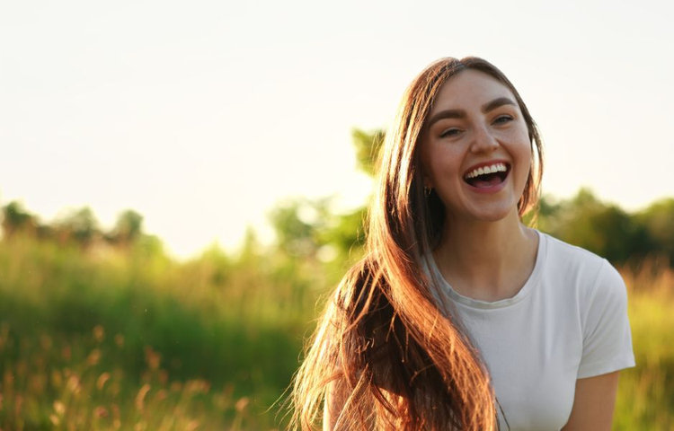 Mladá žena s dlhými hnedými vlasmi sa smeje na slnečnej lúke pri západe slnka, symbolizujúc radosť a zdravú pleť ako výsledok správnej starostlivosti o pleť v máji.